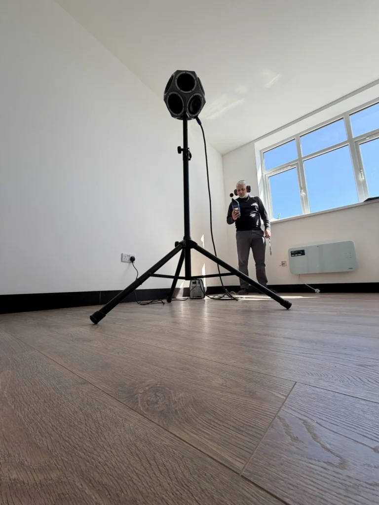 GC Reports professional sound insulation testing on-site. The image shows a technician using a dodecahedron speaker and sound level meter to ensure building compliance and acoustic quality in a modern residential space.